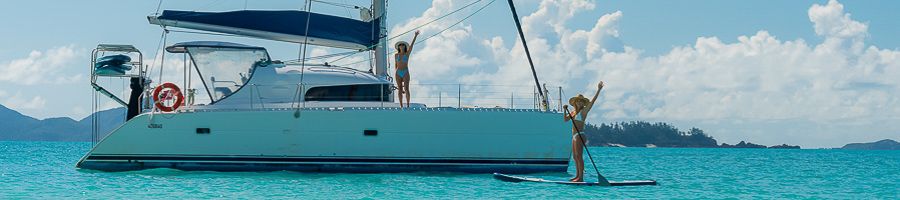 Whitsunday Blue 3D3N A white catamaran yacht anchored in blue water with a girl on a paddle board next to it