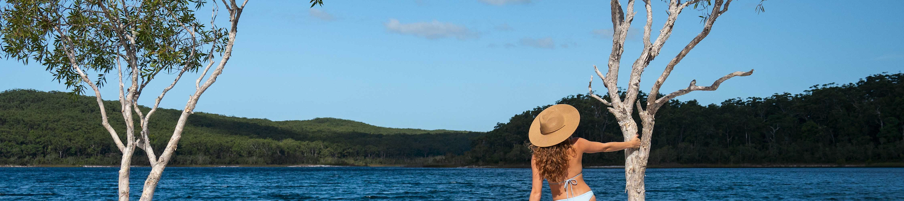 a woman in a brown hat standing in front of a lake holding a tree