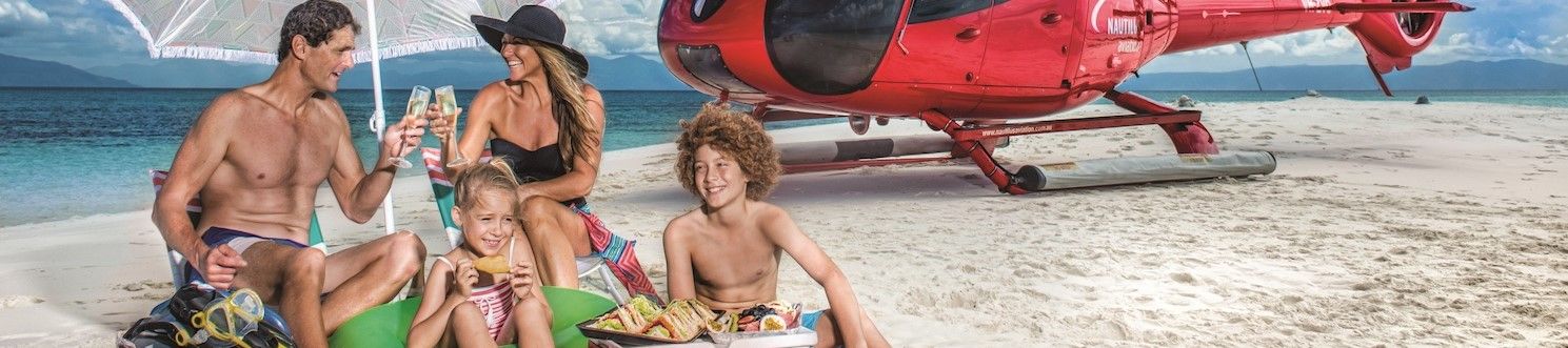 Family of four toast with sparkling wine under a beach umbrella, children enjoy gourmet picnic, helicopter sits on white sands in background