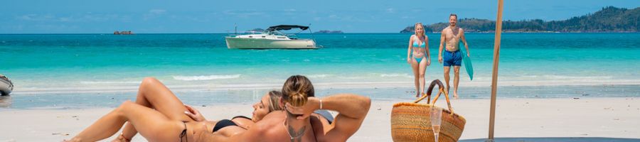 A couple sitting on Whitehaven Beach with a champagne picnic and Salty Yacht in the background